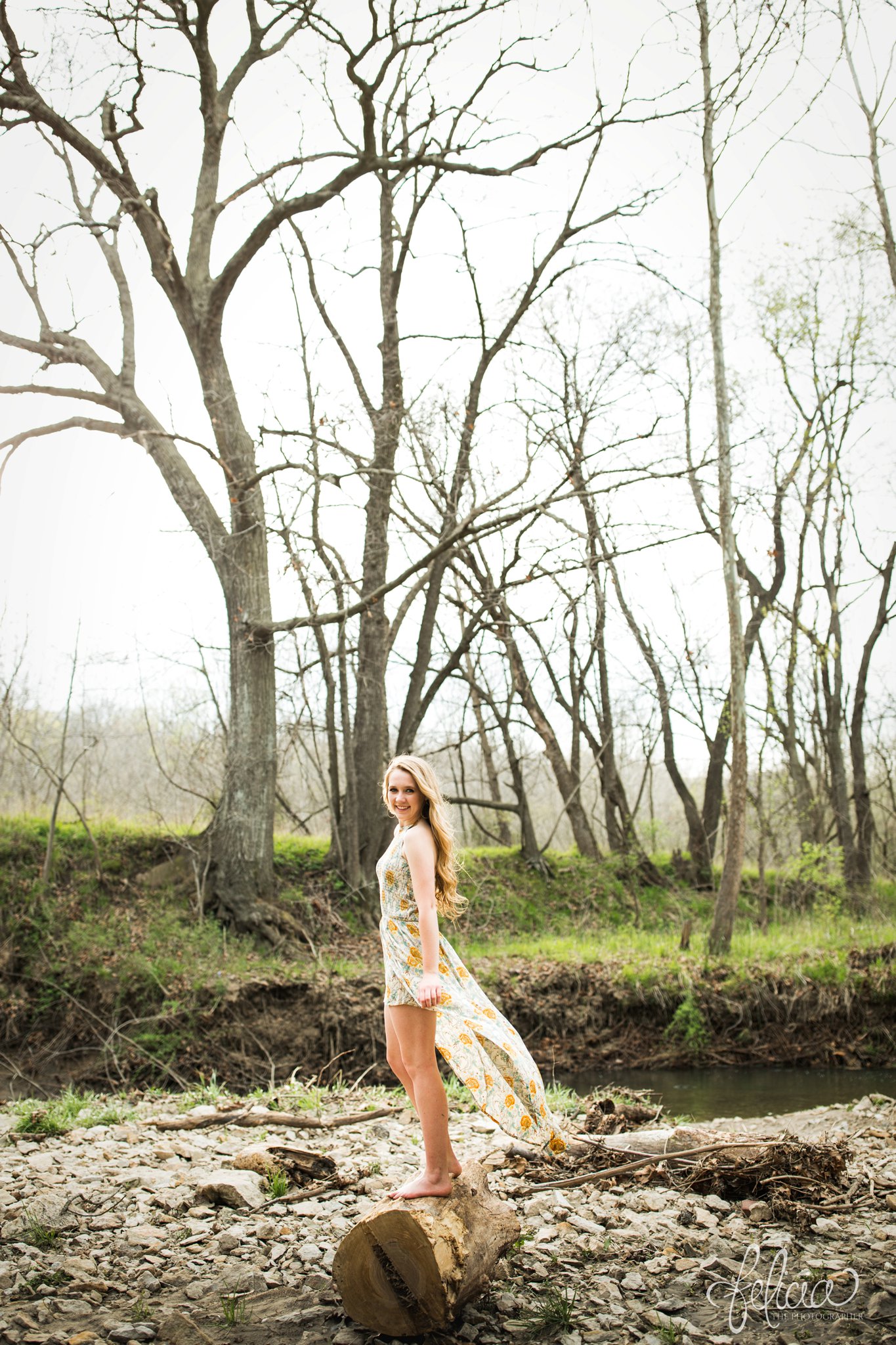 senior pictures | images by feliciathephotographer.com | Kansas City | rustic | long blonde hair | nature background | trees | flower print dress | yellow dress | elegant | dramatic | stream | standing in trees 