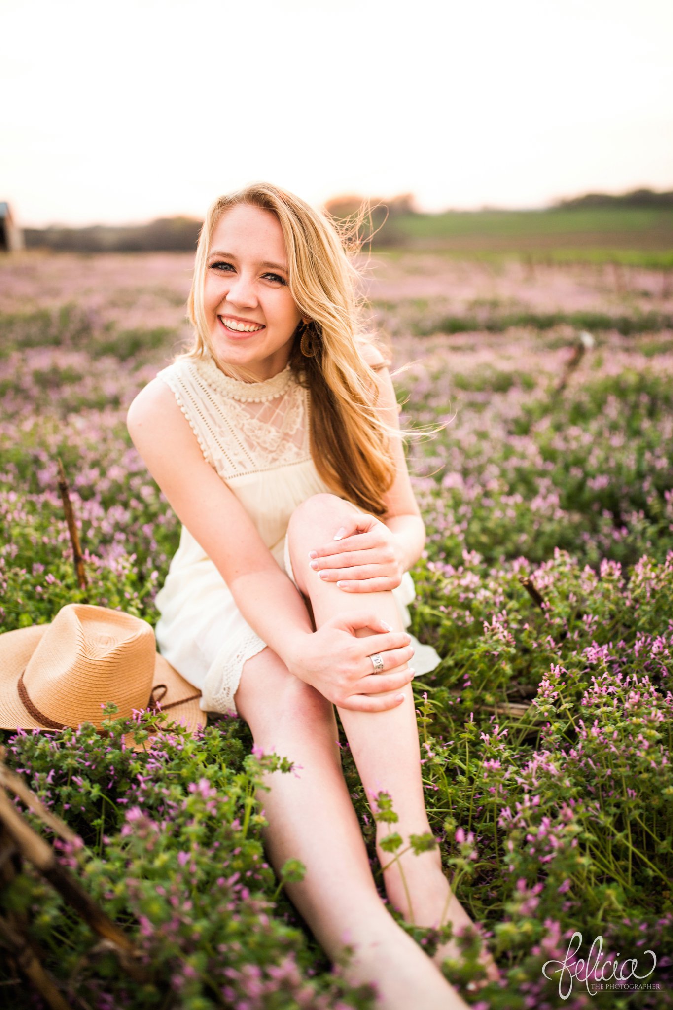 senior pictures | images by feliciathephotographer.com | Kansas City | rustic | long blonde hair | nature background | white dress | elegant | dramatic | candid | dramatic pose | flower field | sitting in flowers | cross legged | purple flowers | blowing kisses 