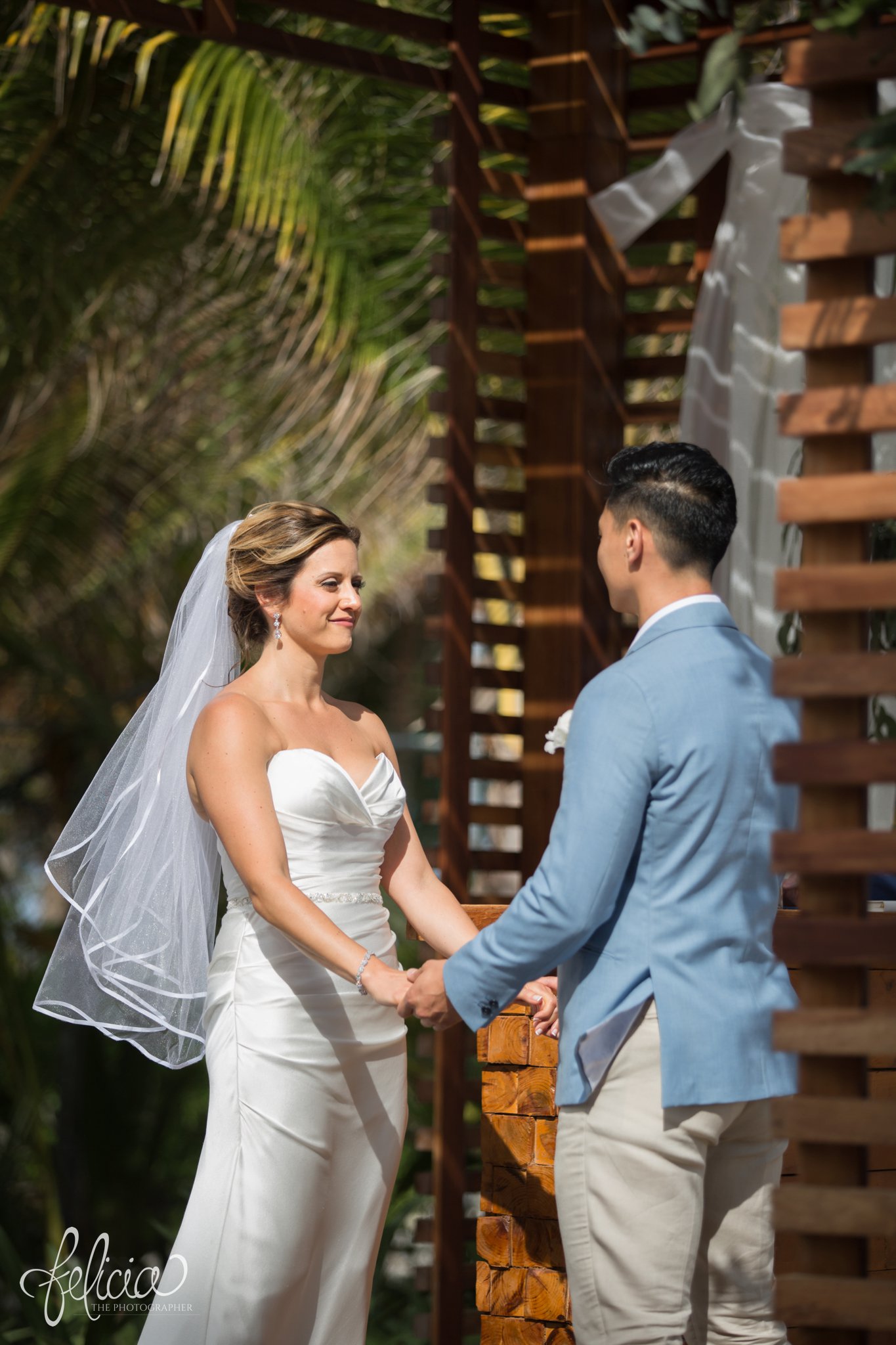 images by feliciathephotographer.com | Destination Beach Wedding | Unicco 20 87 | Photographer | L&S Travel | ceremony | strapless dress | chic | blue jacket | veil | palm trees | stone altar | wooden background | holding hands | exchanging vows 